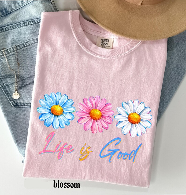 Pode incluir: Camiseta rosa claro com gola redonda e tr&ecirc;s margaridas azuis, rosas e brancas com centros amarelos. O texto "Life is Good" est&aacute; escrito em um script colorido. A palavra "blossom" est&aacute; escrita na parte inferior.