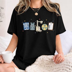 May include: A black short-sleeve t-shirt featuring a graphic print of five cartoon medical imaging items: an X-ray of a rib cage, a lead apron, an X-ray machine, a radiation symbol, and a clipboard, all decorated with small colorful stars.