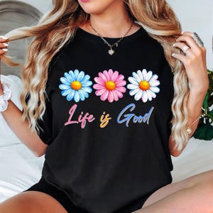 Pode incluir: Camiseta preta com tr&ecirc;s margaridas azuis, rosas e brancas, cada uma com um centro amarelo. Abaixo, a frase "Life is Good" est&aacute; escrita em uma fonte colorida e gradiente. Uma pe&ccedil;a casual para o dia a dia.