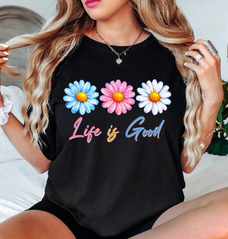 Pode incluir: Camiseta preta com tr&ecirc;s margaridas azuis, rosas e brancas, cada uma com um centro amarelo. Abaixo, a frase "Life is Good" est&aacute; escrita em uma fonte colorida e gradiente. Uma pe&ccedil;a casual para o dia a dia.