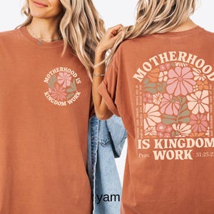 May include: A terracotta-coloured t-shirt with the words "MOTHERHOOD IS KINGDOM WORK" and a floral design. The back of the shirt has the same text and a larger floral arrangement with text from Proverbs 31:25-27.