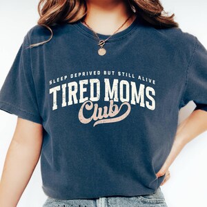 May include: A navy blue short-sleeve t-shirt with the text "SLEEP DEPRIVED BUT STILL ALIVE TIRED MOMS Club" in white and pink lettering. The shirt is a relaxed fit and made of a soft material. The word "midnight" is at the bottom of the shirt.