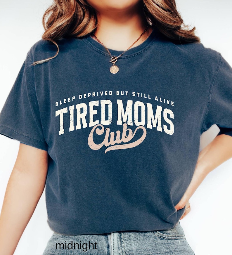 May include: A navy blue short-sleeve t-shirt with the text "SLEEP DEPRIVED BUT STILL ALIVE TIRED MOMS Club" in white and pink lettering. The shirt is a relaxed fit and made of a soft material. The word "midnight" is at the bottom of the shirt.