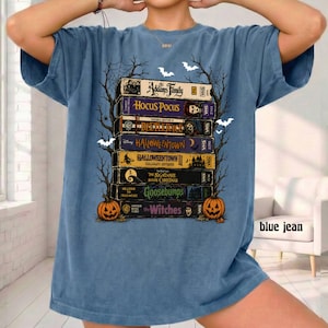 Op de afbeelding: Een blauw t-shirt met een grafische print van een stapel VHS-banden met titels zoals The Addams Family, Hocus Pocus, Beetlejuice, Halloweentown, Halloweentown II, The Nightmare Before Christmas, Goosebumps en The Witches, omlijst door bomen.