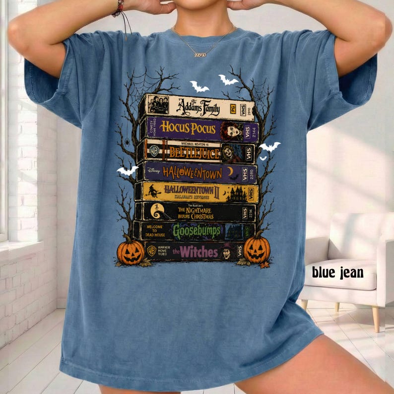 Op de afbeelding: Een blauw t-shirt met een grafische print van een stapel VHS-banden met titels zoals The Addams Family, Hocus Pocus, Beetlejuice, Halloweentown, Halloweentown II, The Nightmare Before Christmas, Goosebumps en The Witches, omlijst door bomen.