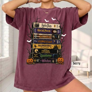 Op de afbeelding: Een bessenkleurig t-shirt met een grafische print van een stapel VHS-banden met titels zoals The Addams Family, Hocus Pocus, Beetlejuice, Halloweentown, The Nightmare Before Christmas, Goosebumps en The Witches, omlijst door boomtakken.