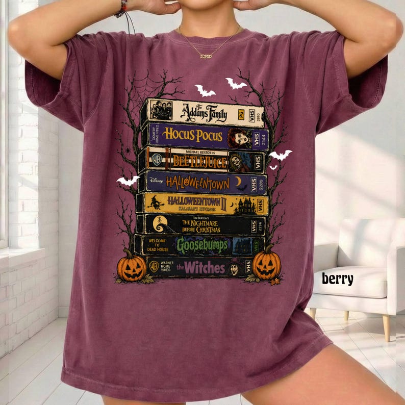 Op de afbeelding: Een bessenkleurig t-shirt met een grafische print van een stapel VHS-banden met titels zoals The Addams Family, Hocus Pocus, Beetlejuice, Halloweentown, The Nightmare Before Christmas, Goosebumps en The Witches, omlijst door boomtakken.