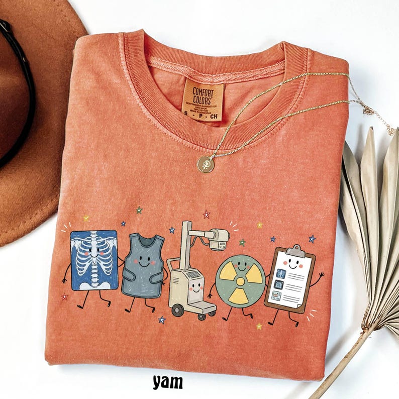 May include: A terracotta-colored t-shirt with a crew neckline and a Comfort Colors label. The graphic features five cartoon characters: an X-ray, a lead apron, an X-ray machine, a radiation symbol, and a clipboard, all accented by small colorful stars.