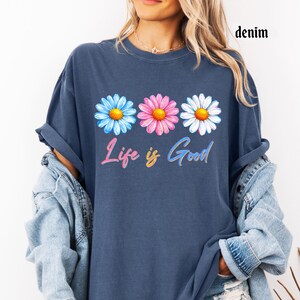 Pode incluir: T-shirt azul denim com um design floral. A camisa apresenta tr&ecirc;s margaridas em azul, rosa e branco, com centros amarelos. A frase "Life is Good" est&aacute; escrita em um script colorido abaixo das flores.