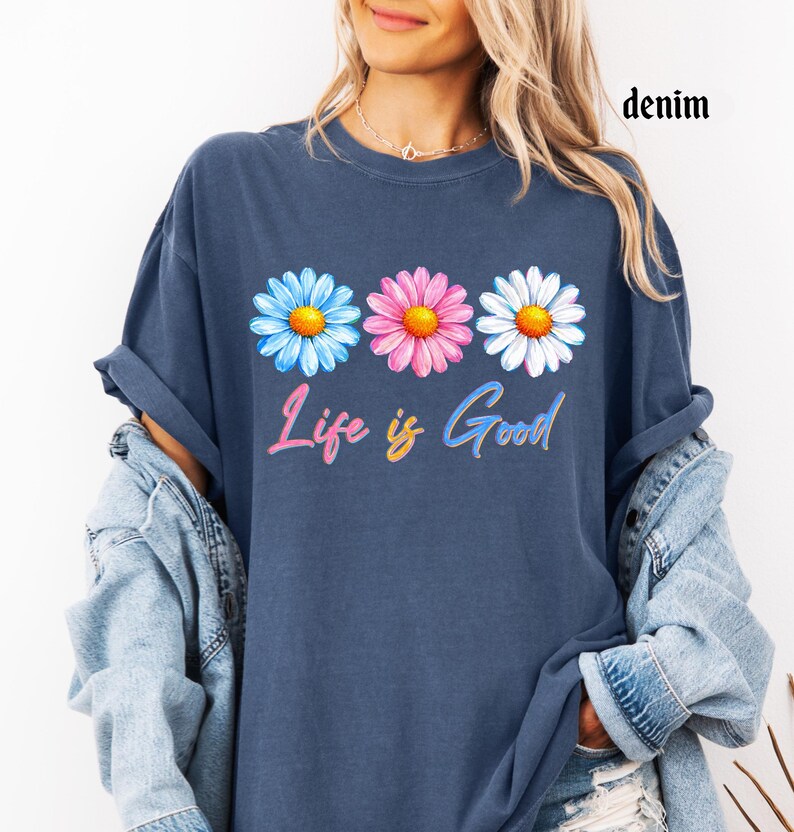 Pode incluir: T-shirt azul denim com um design floral. A camisa apresenta tr&ecirc;s margaridas em azul, rosa e branco, com centros amarelos. A frase "Life is Good" est&aacute; escrita em um script colorido abaixo das flores.