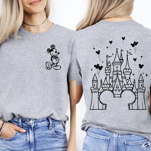 Puede incluir: Una camiseta gris con un dibujo en blanco y negro de Mickey Mouse en la parte delantera y un dise&ntilde;o de castillo en la parte trasera. El dise&ntilde;o del castillo incluye orejas y globos de Mickey Mouse.