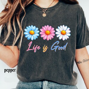 Pode incluir: Camiseta cinza escuro com tr&ecirc;s margaridas azuis, rosas e brancas com centros amarelos. A frase "Life is Good" est&aacute; escrita em letras coloridas abaixo das flores. A palavra "pepper" est&aacute; impressa no canto inferior esquerdo.