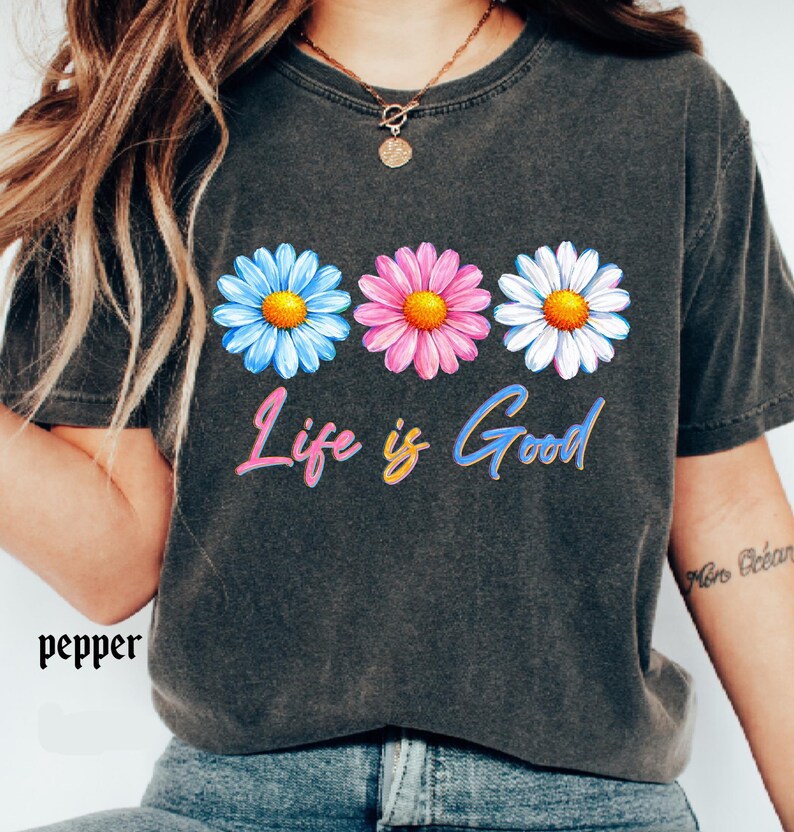Pode incluir: Camiseta cinza escuro com tr&ecirc;s margaridas azuis, rosas e brancas com centros amarelos. A frase "Life is Good" est&aacute; escrita em letras coloridas abaixo das flores. A palavra "pepper" est&aacute; impressa no canto inferior esquerdo.