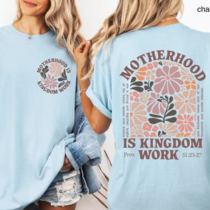 May include: Pale blue t-shirt with the text "MOTHERHOOD IS KINGDOM WORK" and a floral design. The back of the shirt features the same text and a larger floral arrangement. The shirt has rolled-up sleeves.
