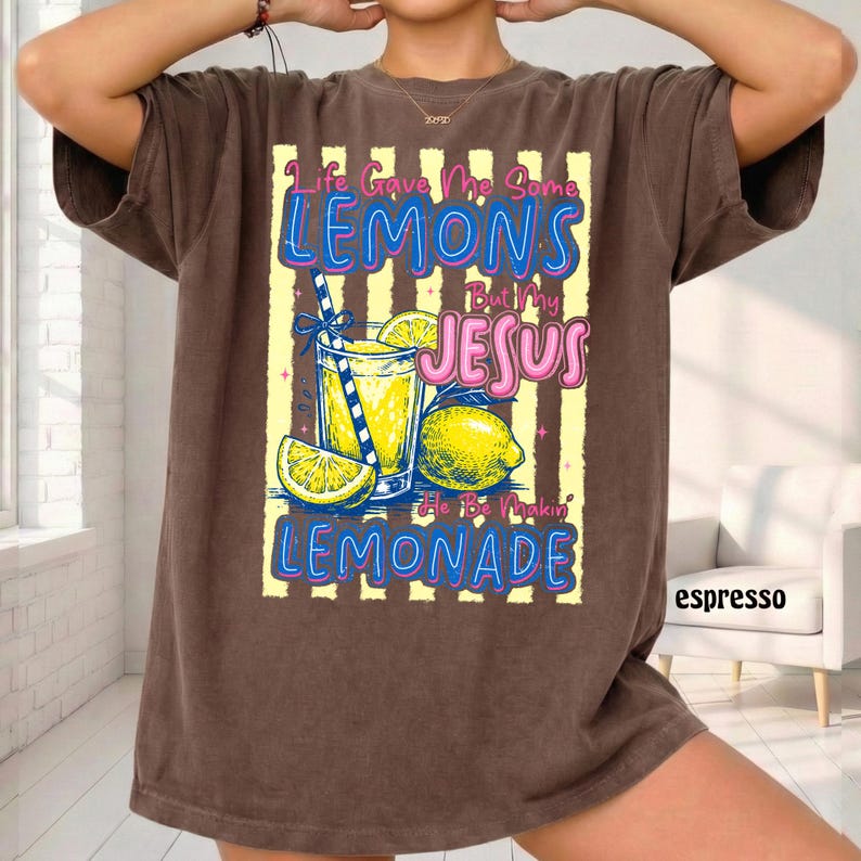 May include: Espresso-colored t-shirt with a graphic of a lemonade drink, lemons, and the text "Life Gave Me Some Lemons But My Jesus He Be Makin' Lemonade" in blue and pink. The shirt has a vintage, worn look.