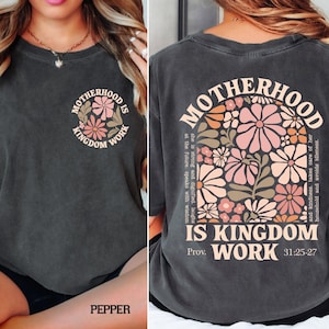 May include: Dark grey t-shirt with the words "MOTHERHOOD IS KINGDOM WORK" and a floral design. The back of the shirt has the same text and a larger floral design with a quote from Proverbs 31:25-27.