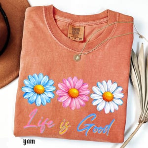 Pode incluir: Uma camiseta coral com tr&ecirc;s flores de margarida em azul, rosa e branco, cada uma com um centro amarelo. As palavras "Life is Good" est&atilde;o escritas em um script colorido abaixo das flores. Um colar de ouro e um chap&eacute;u marrom tamb&eacute;m s&atilde;o vis&iacute;veis.