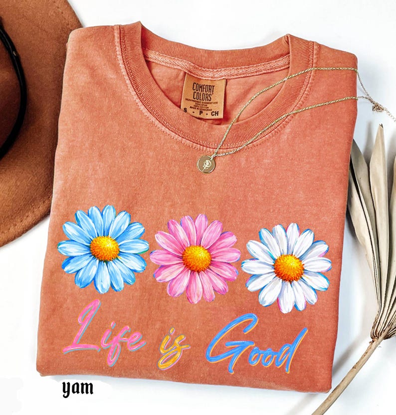 Pode incluir: Uma camiseta coral com tr&ecirc;s flores de margarida em azul, rosa e branco, cada uma com um centro amarelo. As palavras "Life is Good" est&atilde;o escritas em um script colorido abaixo das flores. Um colar de ouro e um chap&eacute;u marrom tamb&eacute;m s&atilde;o vis&iacute;veis.