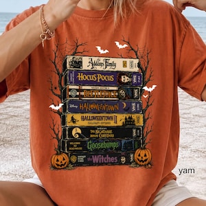 Op de afbeelding: Een oranje t-shirt met een grafische print van een stapel van acht VHS-banden met titels zoals The Addams Family, Hocus Pocus, Beetlejuice, Halloweentown, Halloweentown II, The Nightmare Before Christmas, Goosebumps en The Witches.