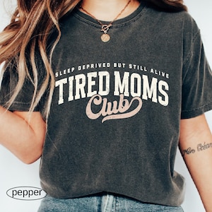 May include: Dark gray t-shirt with the text "SLEEP DEPRIVED BUT STILL ALIVE TIRED MOMS Club" in white and pink lettering. The shirt is a crew neck style and has a vintage, worn look.