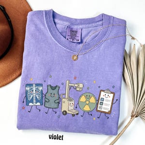 May include: A violet short-sleeve t-shirt with a Comfort Colors label at the neck. The graphic features five cartoon characters: an X-ray, a lead apron, an imaging machine, a radiation symbol, and a clipboard, all accented by small colorful stars.
