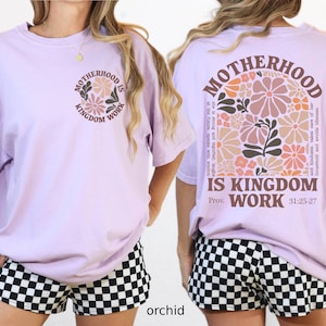 May include: Lilac t-shirt featuring the text "MOTHERHOOD IS KINGDOM WORK" and a floral design on the front and back. The back also includes the Bible verse Proverbs 31:25-27. The model is wearing black and white checkered shorts.