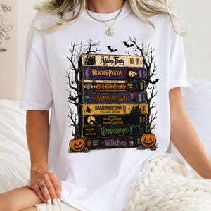 Op de afbeelding: Een wit t-shirt met een grafische print van een stapel VHS-banden met de titels The Addams Family, Hocus Pocus, Beetlejuice, Halloweentown, Halloweentown II, The Nightmare Before Christmas, Goosebumps en The Witches, omlijst door spinnenwebben en pompoenen.
