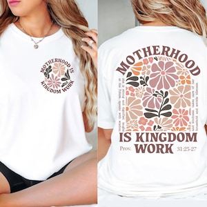 May include: White t-shirt featuring the text "MOTHERHOOD IS KINGDOM WORK" in a circular design on the front and back. The back design includes a floral arrangement in pink, peach, and brown, with a quote from Proverbs 31:25-27.