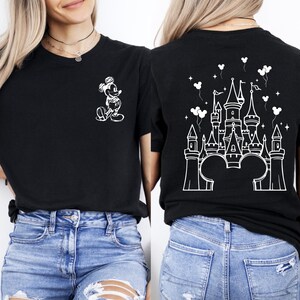 Puede incluir: Camiseta negra con un boceto blanco de Mickey Mouse en la parte delantera y un dise&ntilde;o de castillo en la parte trasera. El dise&ntilde;o del castillo incluye orejas de Mickey Mouse, globos y estrellas. La camiseta se combina con vaqueros azules.