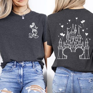 Puede incluir: Camiseta gris oscuro con un boceto blanco de Mickey Mouse en la parte delantera y un dise&ntilde;o de castillo blanco en la parte trasera. El dise&ntilde;o del castillo incluye orejas de Mickey Mouse, globos y estrellas. La camiseta se combina con vaqueros azules.