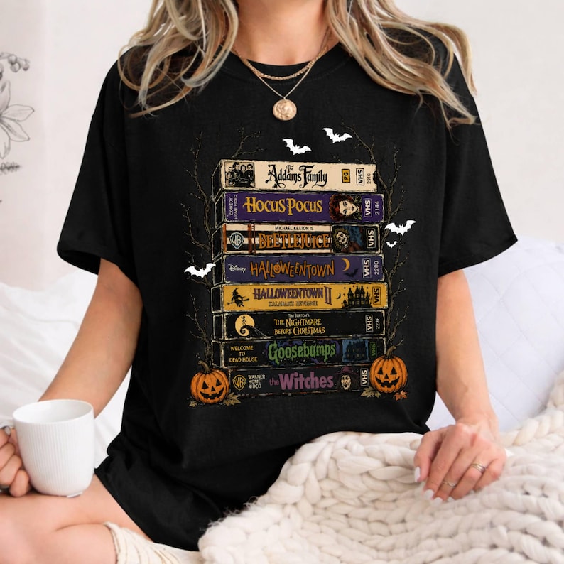 Op de afbeelding: Een zwart T-shirt met korte mouwen en een grafische print van een stapel VHS-banden met titels zoals The Addams Family, Hocus Pocus, Beetlejuice, Halloweentown, Halloweentown II, The Nightmare Before Christmas, Goosebumps en The Witches.