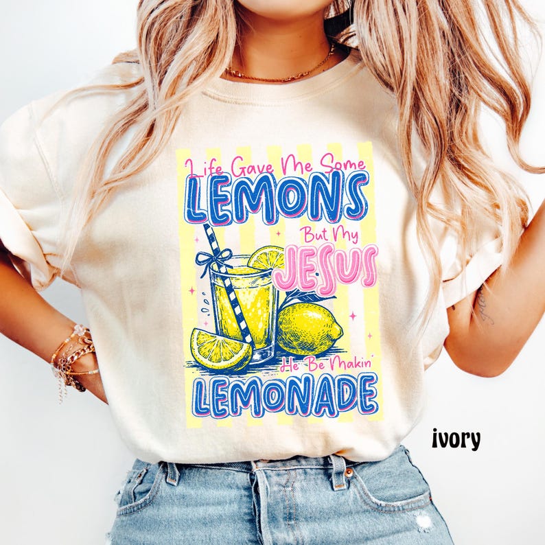 May include: Ivory-colored t-shirt with a lemonade-themed graphic. The design features a glass of lemonade, lemons, and the text "Life Gave Me Some Lemons But My Jesus He Be Makin' Lemonade" in blue and pink.