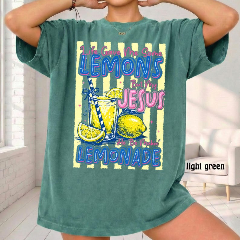 May include: Light green t-shirt with a graphic of a glass of lemonade, lemons, and the text "Life Gave Me Some Lemons But My Jesus Is Makin' Lemonade". The design is in blue, yellow, and pink.