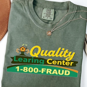 May include: A moss green t-shirt with the words "Quality Learning Center" and "1-800-FRAUD" in a graphic design. A gold-colored necklace with a small pendant rests on the shirt. The shirt is folded.