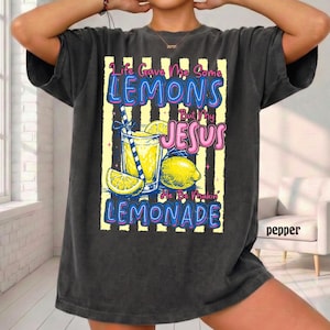 May include: Dark gray t-shirt with a graphic of a glass of lemonade, lemons, and the text "Life Gave Me Some Lemons But My Jesus He Be Makin' Lemonade". The design is in blue, yellow, and pink.