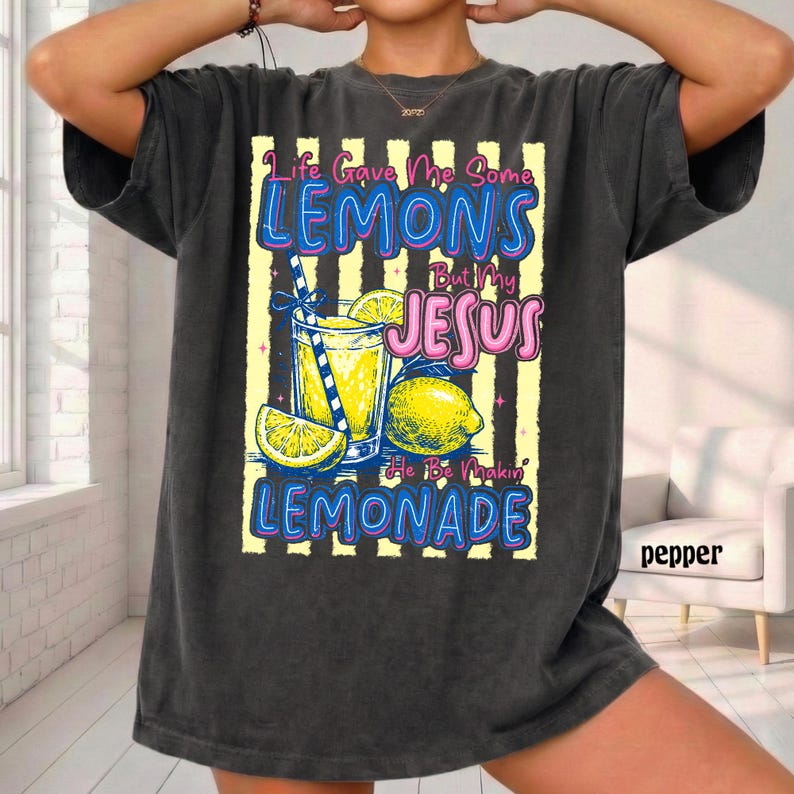 May include: Dark gray t-shirt with a graphic of a glass of lemonade, lemons, and the text "Life Gave Me Some Lemons But My Jesus He Be Makin' Lemonade". The design is in blue, yellow, and pink.