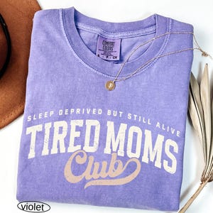 May include: A violet Comfort Colors t-shirt with the text "SLEEP DEPRIVED BUT STILL ALIVE TIRED MOMS Club" in white and tan lettering. A gold necklace with a circular pendant rests on the shirt. The shirt is folded.