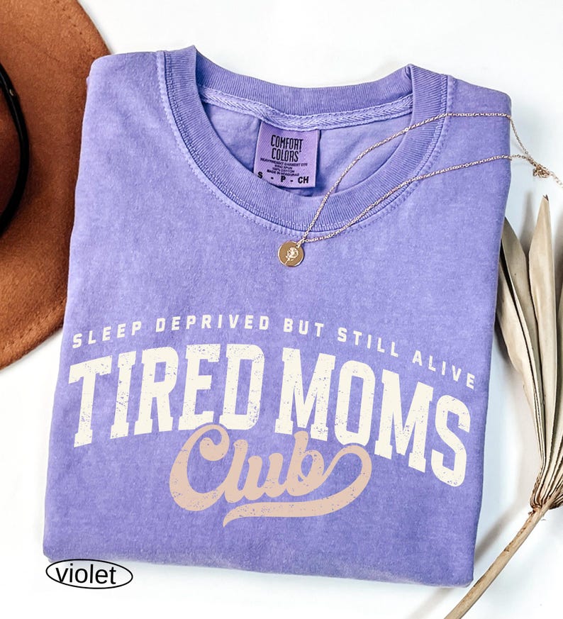 May include: A violet Comfort Colors t-shirt with the text "SLEEP DEPRIVED BUT STILL ALIVE TIRED MOMS Club" in white and tan lettering. A gold necklace with a circular pendant rests on the shirt. The shirt is folded.