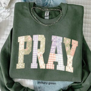 May include: Military green sweatshirt with the word "PRAY" in a patchwork design. The letters are made of floral, gingham, and solid fabric. A silver chain necklace rests on the collar. The sweatshirt is folded on top of a pair of blue jeans.