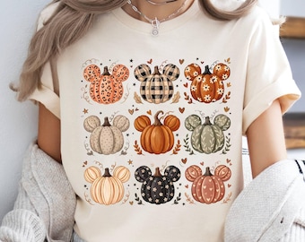 Puede incluir: Una camiseta de manga corta color crema con un estampado gráfico de nueve calabazas dispuestas en cuadrícula. Cada calabaza tiene forma de orejas de ratón y muestra varios patrones, incluyendo estampado de leopardo, cuadros, flores y lunares.