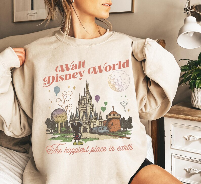 May include: Beige sweatshirt with a vintage-style graphic of Walt Disney World. The design includes the castle, Mickey Mouse, balloons, and the text "Walt Disney World" and "The happiest place in earth".