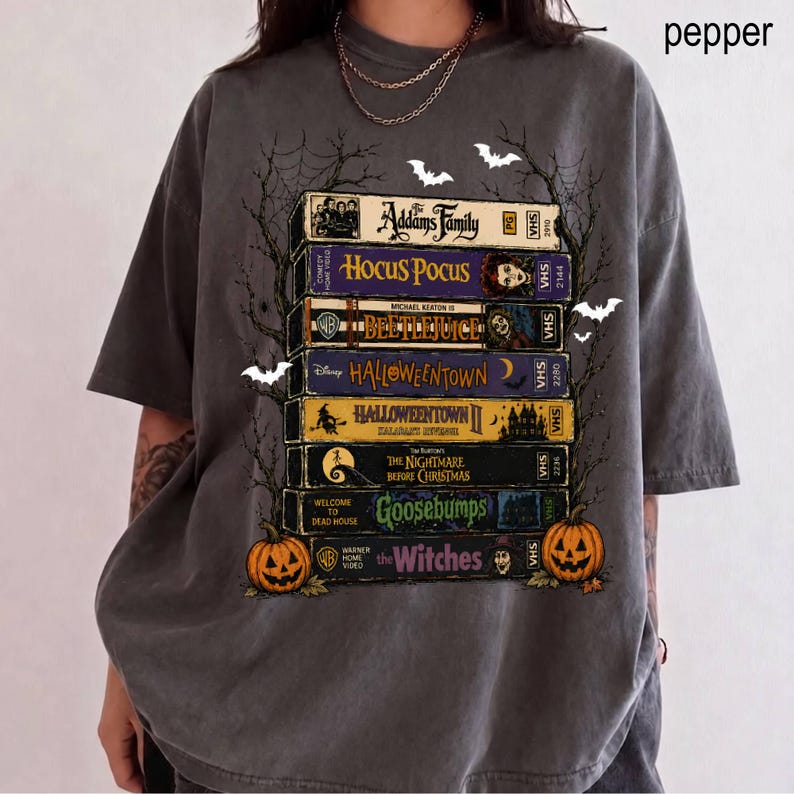 Op de afbeelding: Een antracietgrijs t-shirt met een grafische print van een stapel van zeven VHS-banden met de titels The Addams Family, Hocus Pocus, Beetlejuice, Halloweentown, Halloweentown II, The Nightmare Before Christmas, Goosebumps en The Witches.