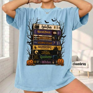 Op de afbeelding: Een lichtblauw t-shirt met een grafische print van een stapel VHS-banden met titels zoals The Addams Family, Hocus Pocus, Beetlejuice, Halloweentown, Halloweentown II, The Nightmare Before Christmas, Goosebumps en The Witches.