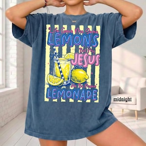 May include: A navy blue t-shirt with the text "Life Gave Me Some Lemons But My Jesus He Be Makin' Lemonade." The graphic includes a glass of lemonade, lemons, and a straw. The shirt is a relaxed fit.