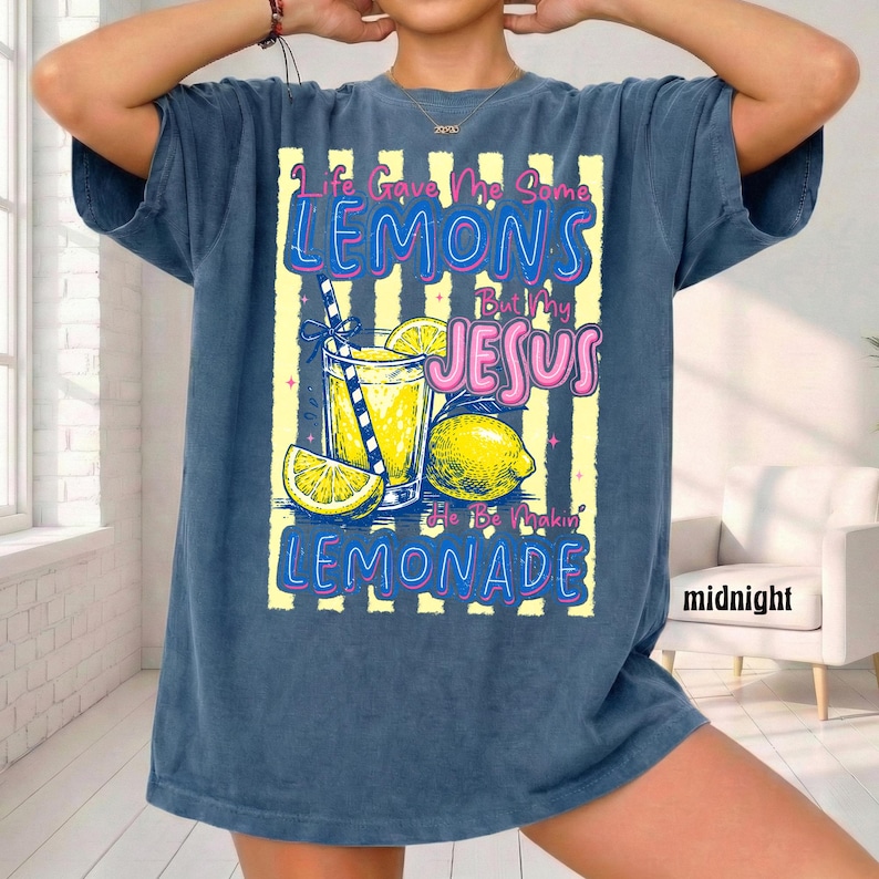May include: A navy blue t-shirt with the text "Life Gave Me Some Lemons But My Jesus He Be Makin' Lemonade." The graphic includes a glass of lemonade, lemons, and a straw. The shirt is a relaxed fit.