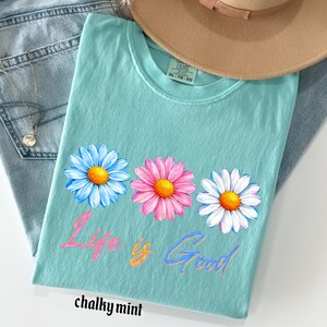 Pode incluir: Uma camiseta turquesa com um design floral de margaridas em azul, rosa e branco, com centros amarelos. A frase "Life is Good" est&aacute; escrita em um script colorido abaixo das flores. A camiseta &eacute; rotulada como "chalky mint".