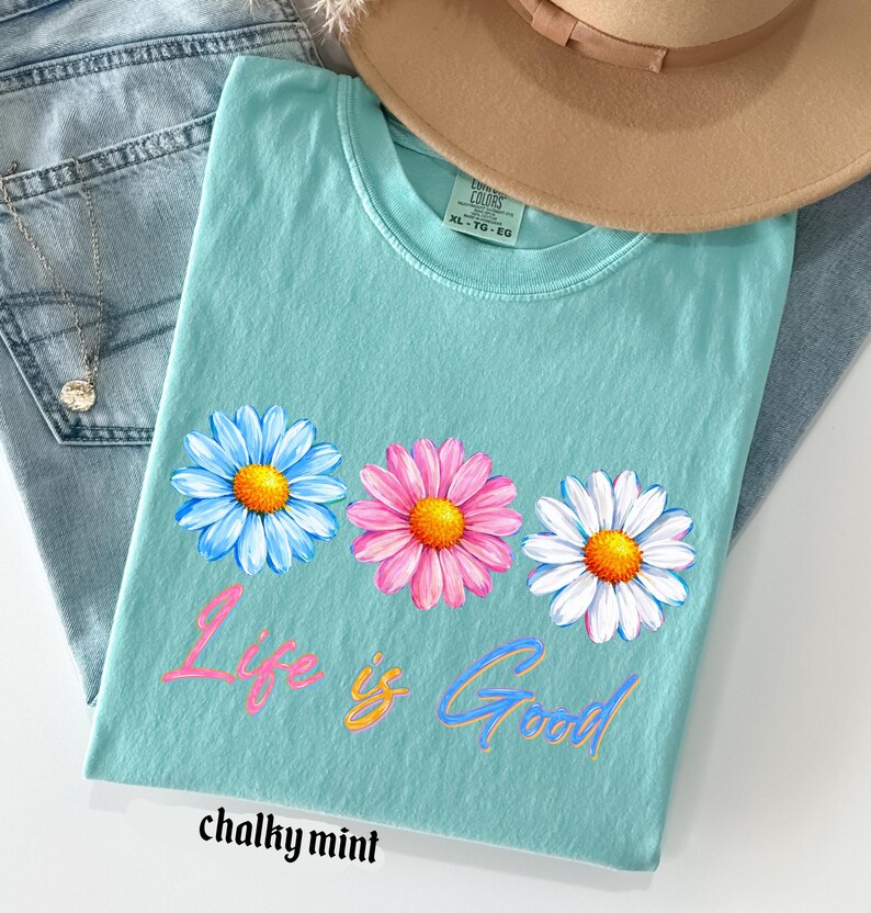 Pode incluir: Uma camiseta turquesa com um design floral de margaridas em azul, rosa e branco, com centros amarelos. A frase "Life is Good" est&aacute; escrita em um script colorido abaixo das flores. A camiseta &eacute; rotulada como "chalky mint".