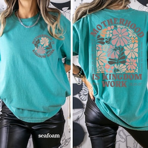 May include: Seafoam green t-shirt featuring the text "MOTHERHOOD IS KINGDOM WORK" and a floral design. The back of the shirt has the same text and a larger floral design. The t-shirt has a relaxed fit.