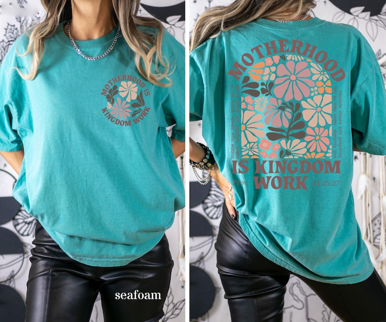 May include: Seafoam green t-shirt featuring the text "MOTHERHOOD IS KINGDOM WORK" and a floral design. The back of the shirt has the same text and a larger floral design. The t-shirt has a relaxed fit.