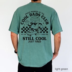 Pu&ograve; includere: T-shirt verde chiaro con la scritta "PROUD MEMBER OF THE COOL DADS CLUB" e una tazza di caff&egrave; dei cartoni animati su uno skateboard. La maglietta dice anche "ALL DAY EVERY DAY STILL COOL JUST TIRED".